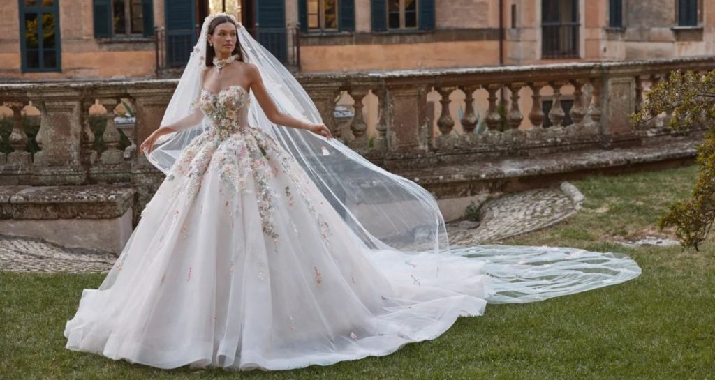 Bride in a floral appliqué ball gown with a long veil standing on grass in front of a historic stone building.