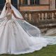 Bride in a floral appliqué ball gown with a long veil standing on grass in front of a historic stone building.