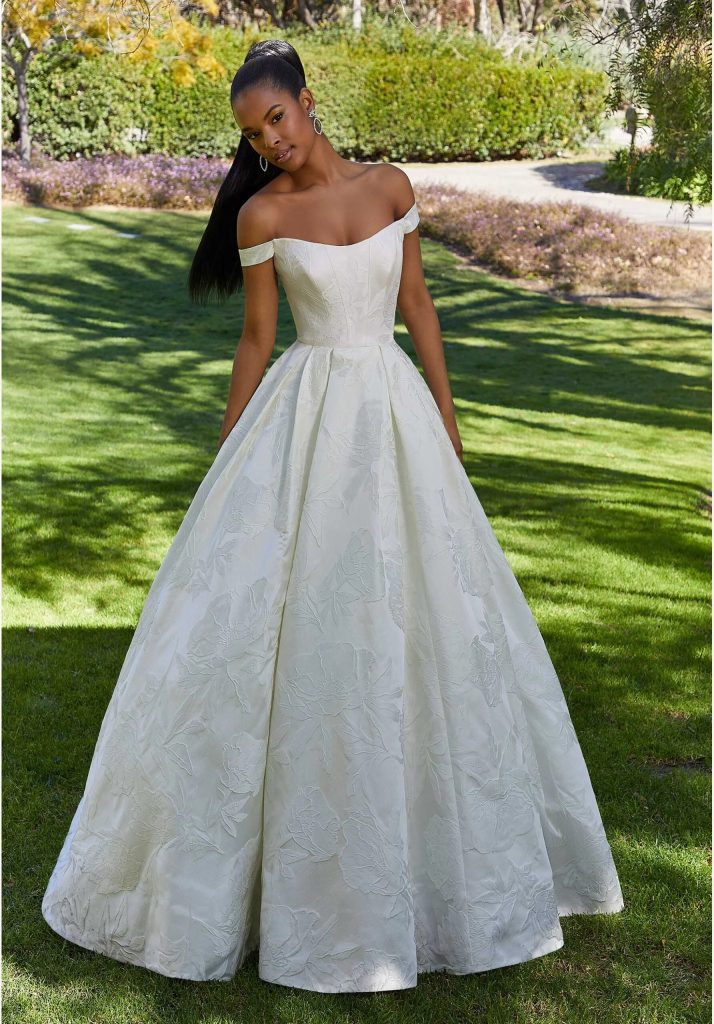 A bride in an off-the-shoulder floral jacquard ball gown stands on a green lawn in a sunny garden setting.