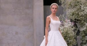 A bride in a square neckline wedding gown with a full skirt stands beside airy white floral arrangements against a stone wall.