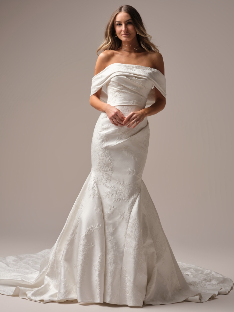 A bride in an off-the-shoulder fitted jacquard wedding gown poses in a studio with a soft neutral background and long train.