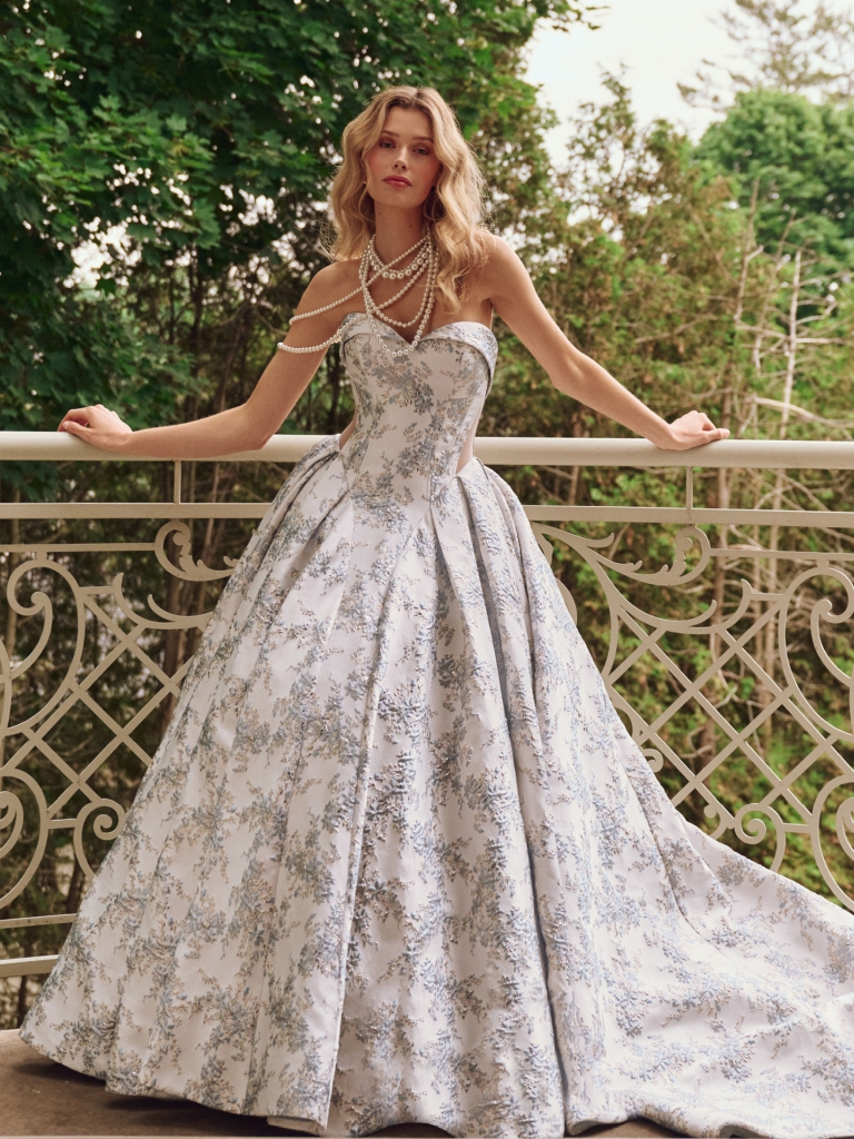 A bride in a strapless silver floral ball gown poses on a balcony with layered pearl necklaces and trees in the background.