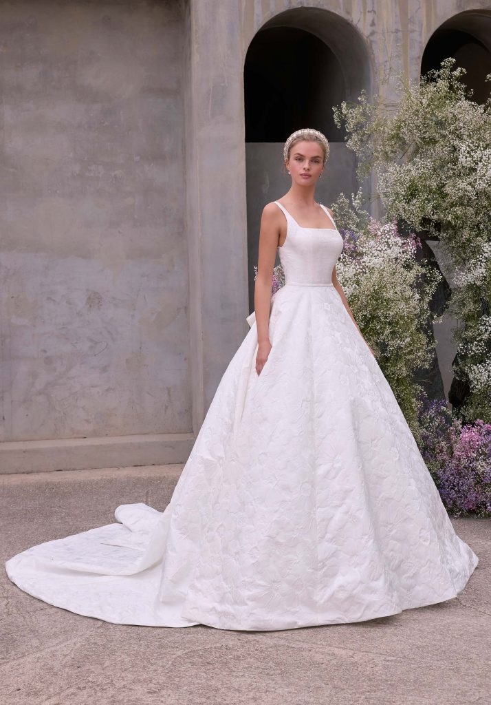 A bride in a square neckline ball gown with a long train stands beside soft white and lavender floral arrangements in a stone courtyard.