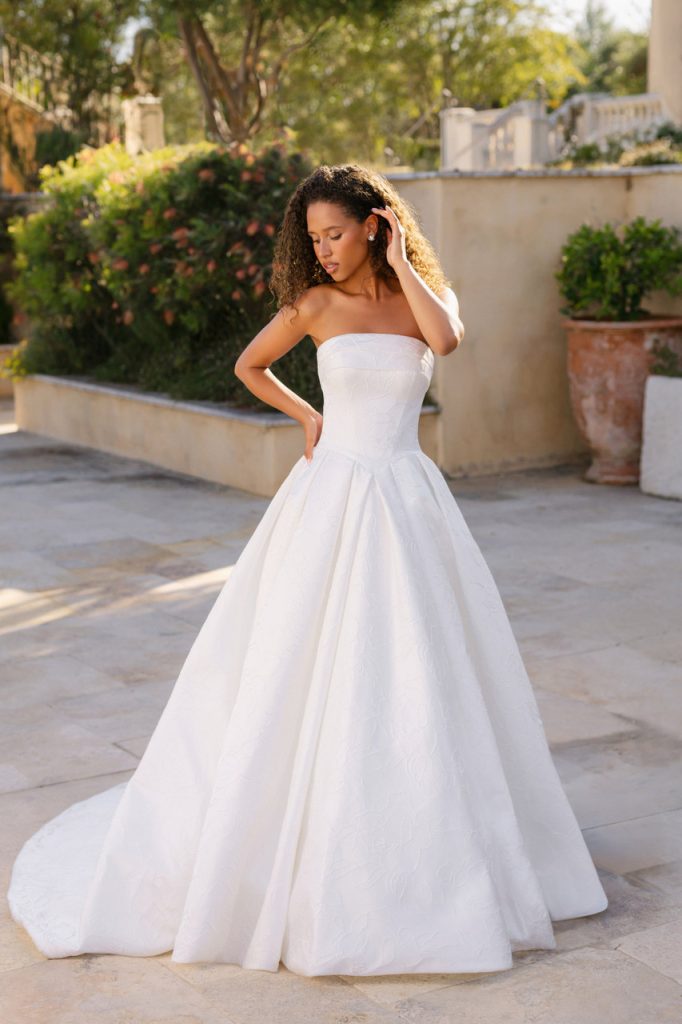 A bride in a strapless A-line wedding gown poses outdoors on a sunlit stone patio surrounded by greenery and garden walls.