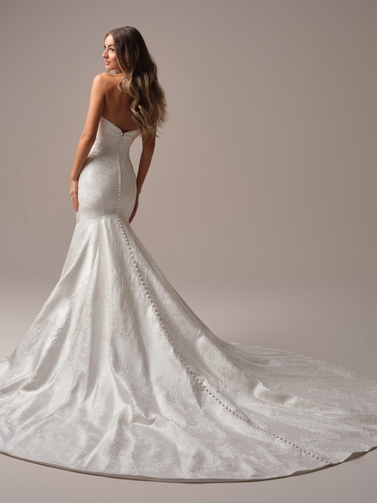 A bride in a fitted strapless jacquard mermaid gown stands with her back turned to show the long train and button detail in a studio.