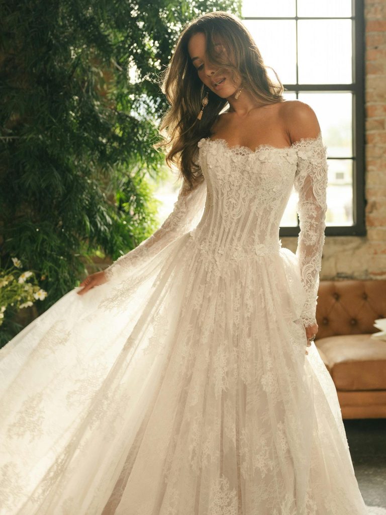 Bride in an off-the-shoulder lace gown stands indoors near large windows with greenery visible, gently holding her skirt.