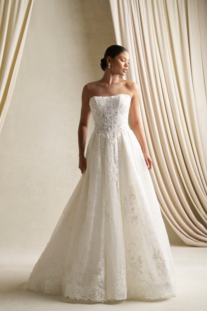 Bride in a strapless lace wedding gown with a full skirt stands in a studio with neutral backdrop and draped fabric.