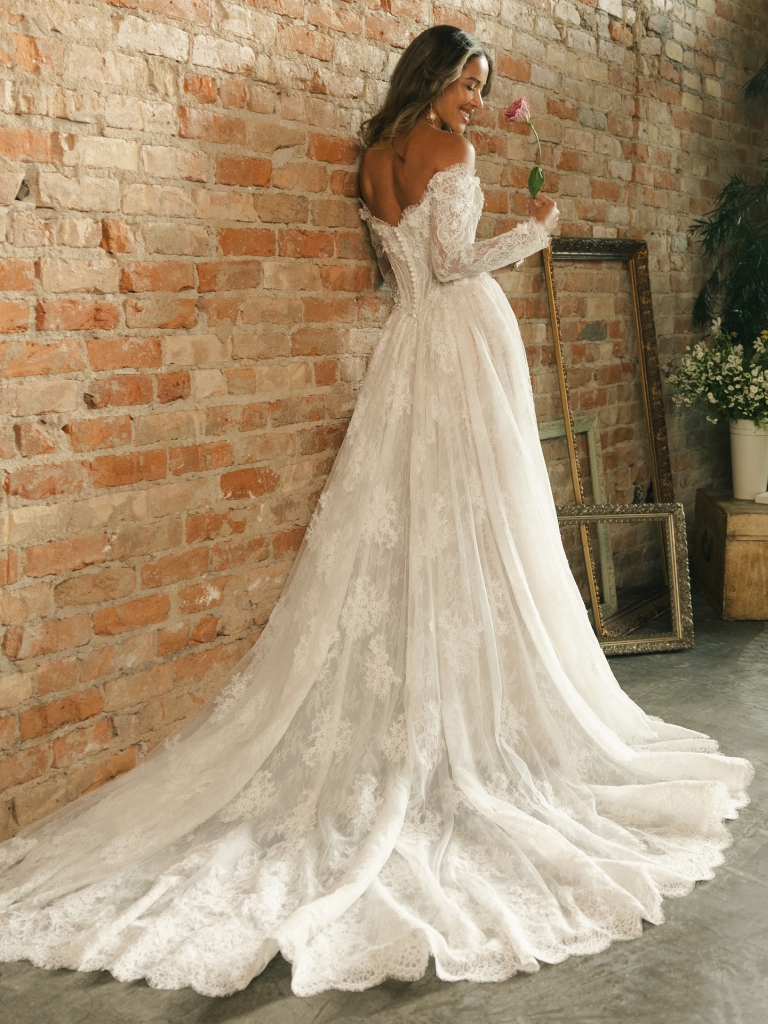 Bride in a long sleeve lace wedding gown stands against a brick wall holding a flower, with a long lace train flowing behind her.
