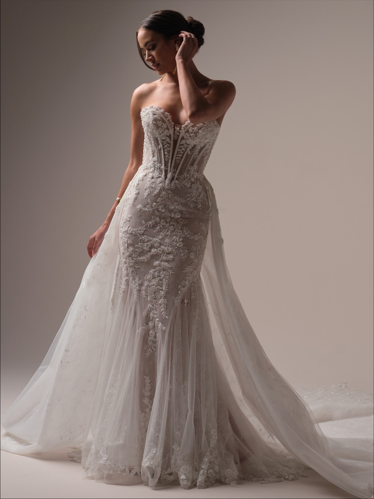 Bride in a fitted lace wedding gown with a strapless neckline and sheer overskirt stands in a studio with a neutral background.
