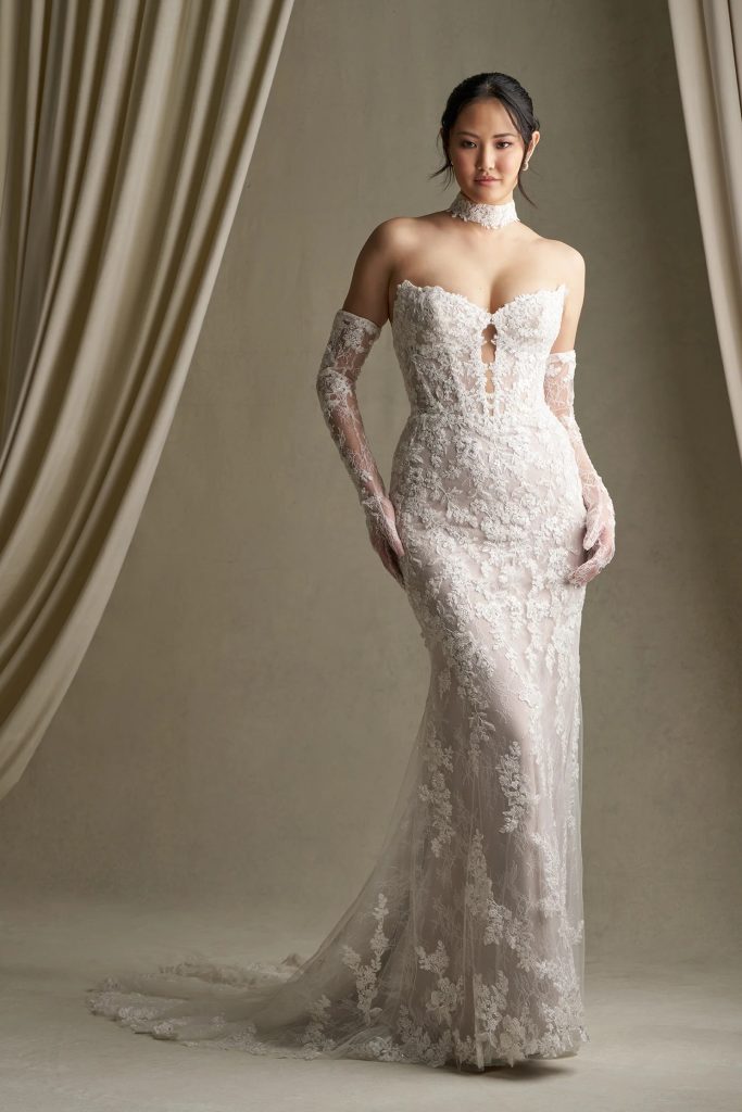 Bride in a strapless lace gown with sheer lace gloves and a choker neckline stands in front of soft draped curtains.