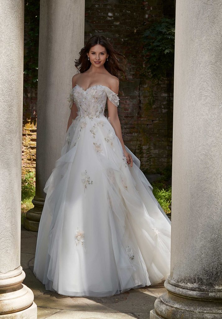 Bride in a voluminous off-the-shoulder wedding dress with floral appliqués stands between stone columns in an outdoor garden.