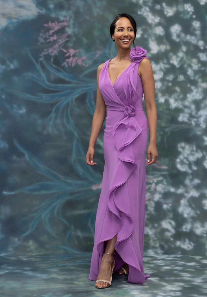 A woman wearing a sleeveless lavender evening gown with floral appliqués and cascading ruffles stands smiling in front of a soft floral backdrop.