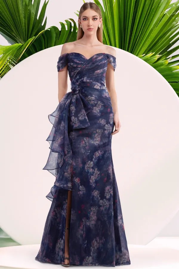 A woman wears a navy off-the-shoulder floral gown with a sweetheart neckline and ruffled side detail in front of tropical leaf accents.