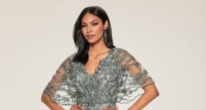 A woman wears a sage green embroidered gown with sheer flutter sleeves and beaded detailing against a neutral background.