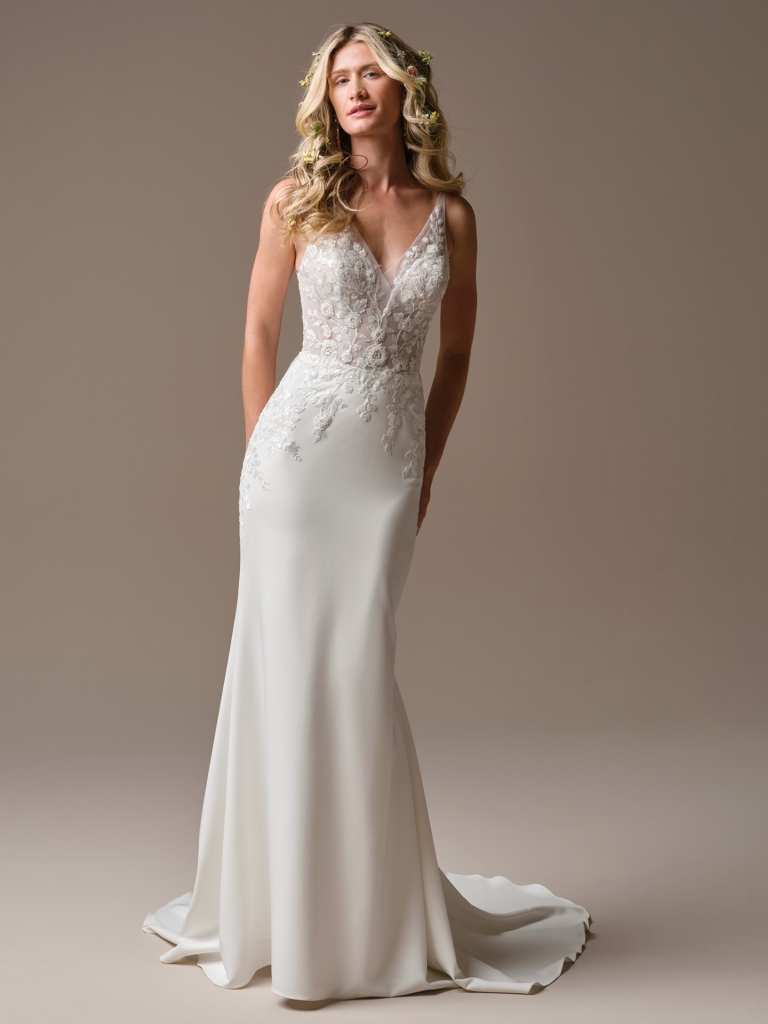 Bride wearing a fitted V-neck wedding gown with floral lace appliqué and a flowing train against a neutral studio background.