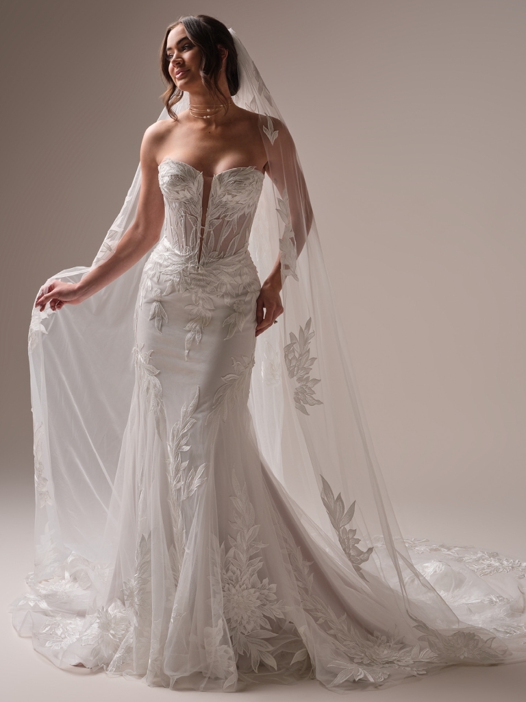 Bride in a fitted lace wedding gown with a plunging neckline and long veil, standing in a studio with soft lighting.