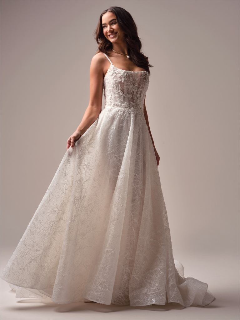 Bride smiling in a delicate lace A-line wedding dress with thin straps, holding the skirt in a studio setting.