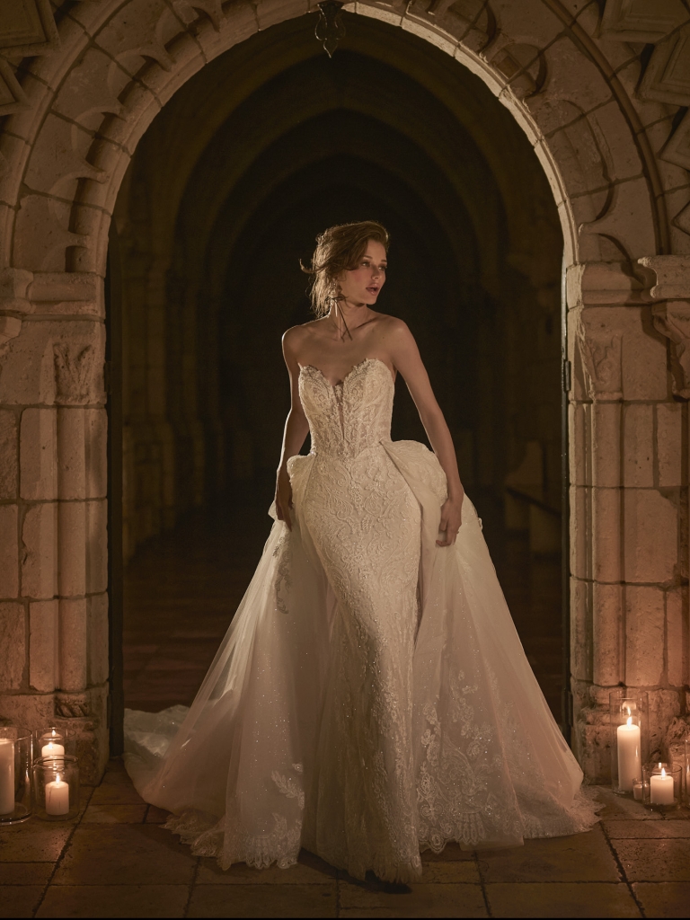 Bride in a fitted lace wedding gown with a sweetheart neckline and dramatic overskirt, standing in a candlelit stone archway.