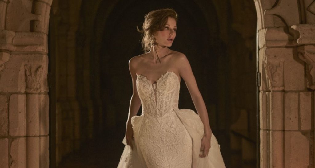 Bride in a strapless lace wedding gown with a sweetheart neckline and detachable overskirt, posing beneath a stone arch.