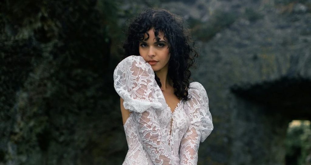 Woman with curly dark hair posing in a fitted lace wedding dress with puff sleeves in front of rocky ruins.