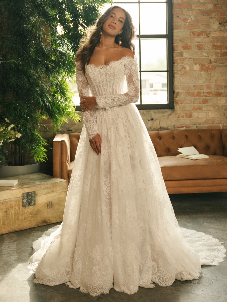 Bride in an off-the-shoulder lace wedding dress with long sleeves, standing in a bright room with a brick wall and window.
