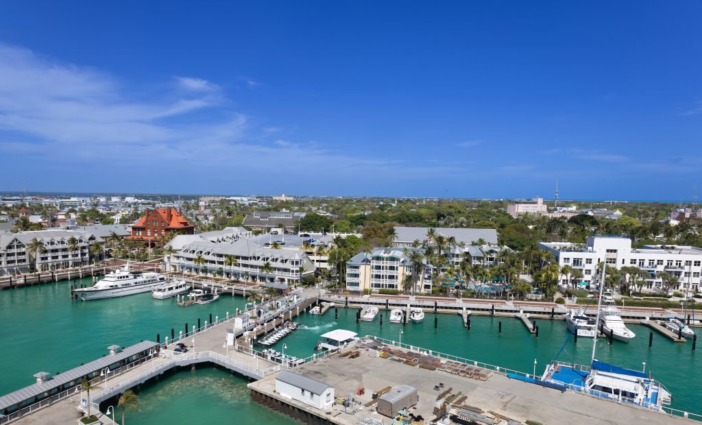 Aerial view of a marina with boats, waterfront hotels, docks, and turquoise water extending toward a coastal cityscape.