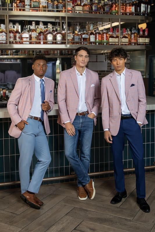 Three men modeling pink plaid sport coats with dress shirts and contrasting trousers inside a bar with shelves of bottles in the background.
