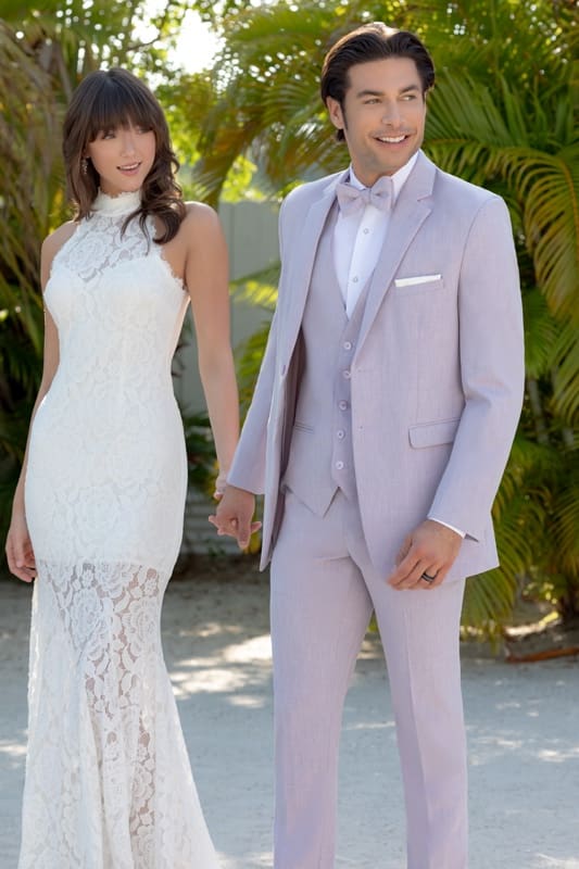 Man wearing a light lavender three-piece suit and bow tie walking hand in hand with a woman in a white lace dress among tropical greenery.