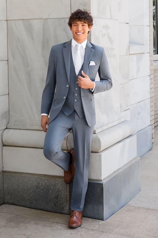 Young man wearing a light blue three-piece suit and tie posing against a stone building exterior.