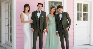 Two men in dark green suits standing with two women in light green dresses in front of a pink wooden building.