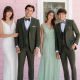 Two men in dark green suits standing with two women in light green dresses in front of a pink wooden building.
