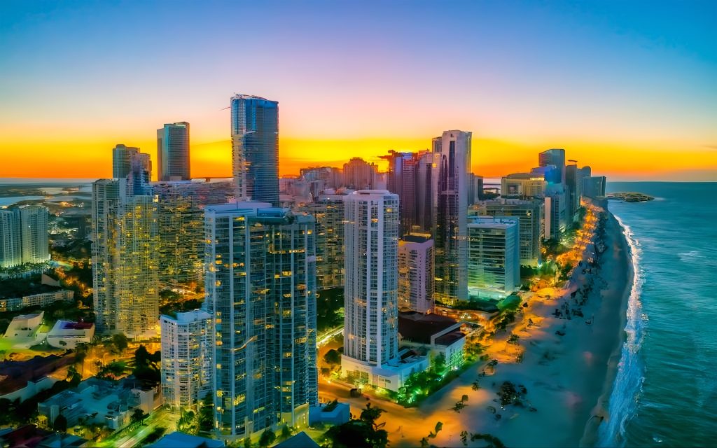 Aerial view of a coastal city skyline at sunset with high-rise buildings lining a beach beside the ocean.