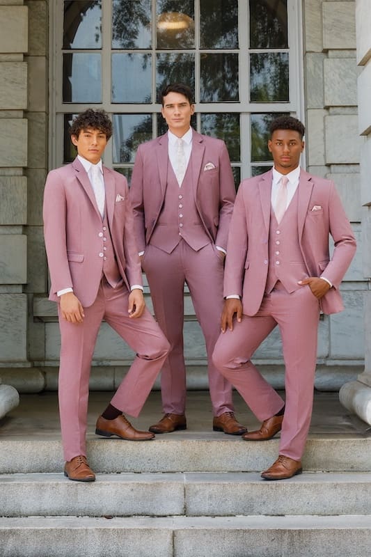 Three men wearing matching dusty rose three-piece suits posing on stone steps in front of a large windowed building entrance.