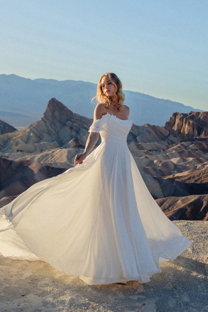 A bride wearing an off-the-shoulder flowing wedding gown poses on a rocky desert overlook with mountains and dramatic rock formations in the background.