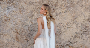 A bride in a sleeveless white wedding gown with long flowing scarf accents stands in profile in front of a textured stone wall.