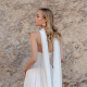 A bride in a sleeveless white wedding gown with long flowing scarf accents stands in profile in front of a textured stone wall.