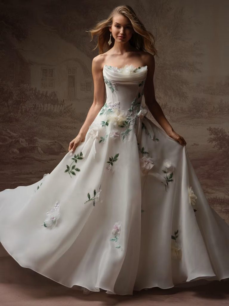 A bride models a strapless white ball gown adorned with floral appliqués while standing against a softly painted studio backdrop.