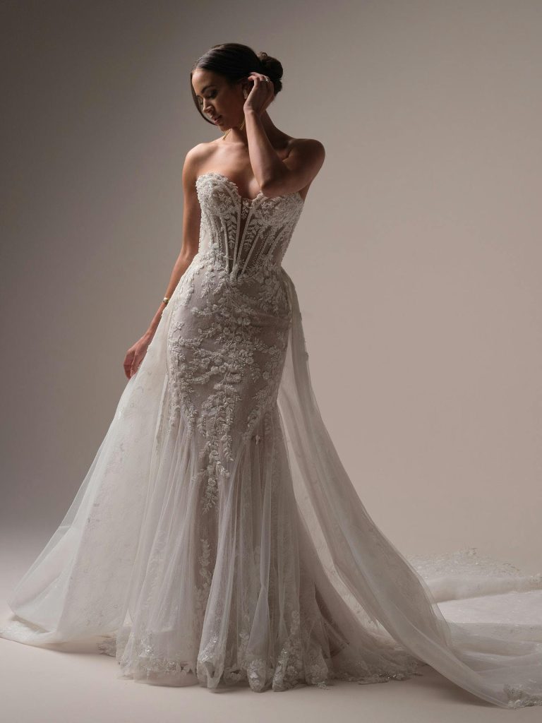 A bride poses in a strapless lace wedding gown with a sheer overskirt and dramatic cathedral-length train against a neutral studio backdrop.