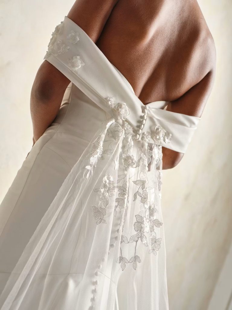 Close-up of the back of an off-the-shoulder satin wedding dress with floral appliqués, covered buttons, and a flowing lace veil.