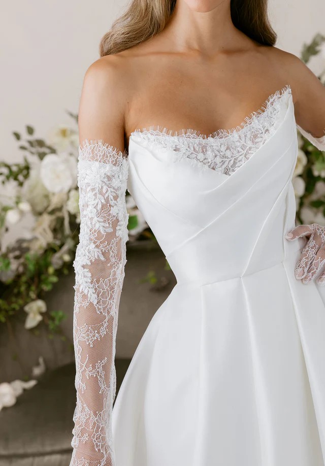 Close-up of a wedding dress with an off-the-shoulder neckline, lace long sleeves, and delicate floral lace trim along the bodice.