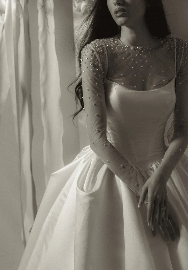 Black-and-white portrait of a bride wearing a satin wedding dress with a sheer pearl-embellished long-sleeve overlay and a full skirt.