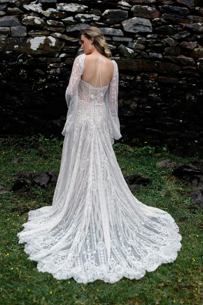 A bride stands with her back turned, showcasing the open-back design and intricate lace detailing of her long-sleeved wedding gown in front of a rustic stone wall.