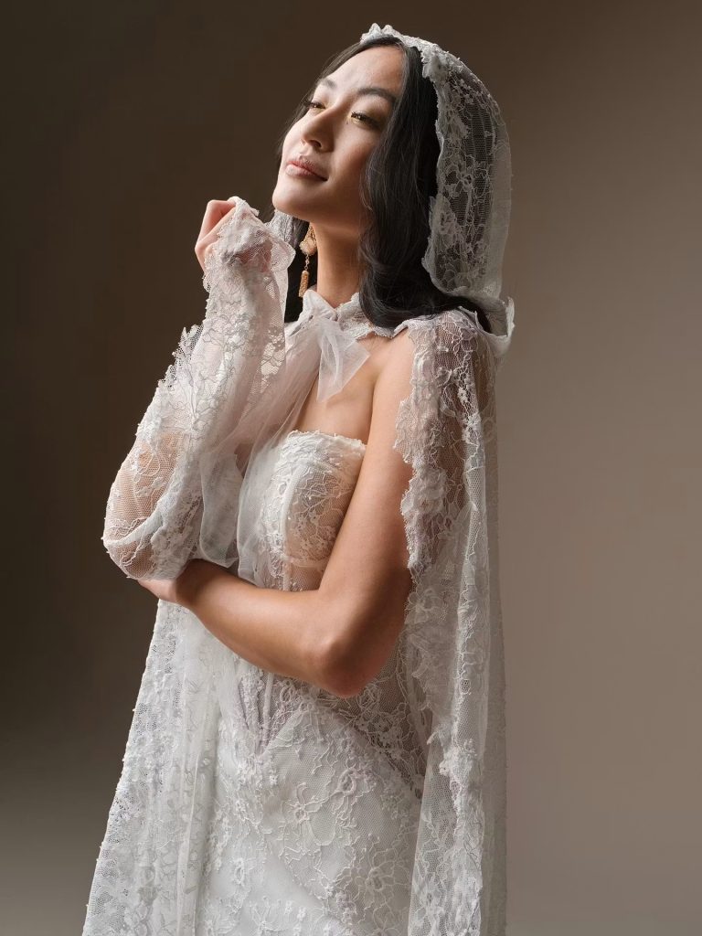 A bride wearing a fitted lace wedding gown with a matching lace hooded cape smiles softly while posing against a neutral studio background.