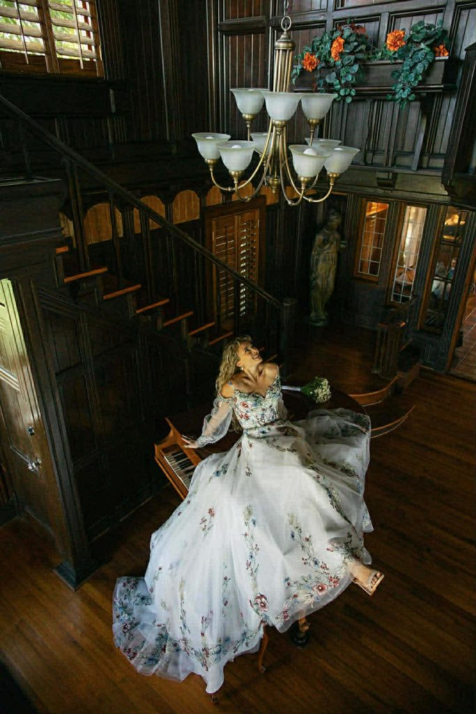 Bride in a floral embroidered wedding dress reclining on a grand piano inside a dark wood historic estate with a sweeping staircase.
