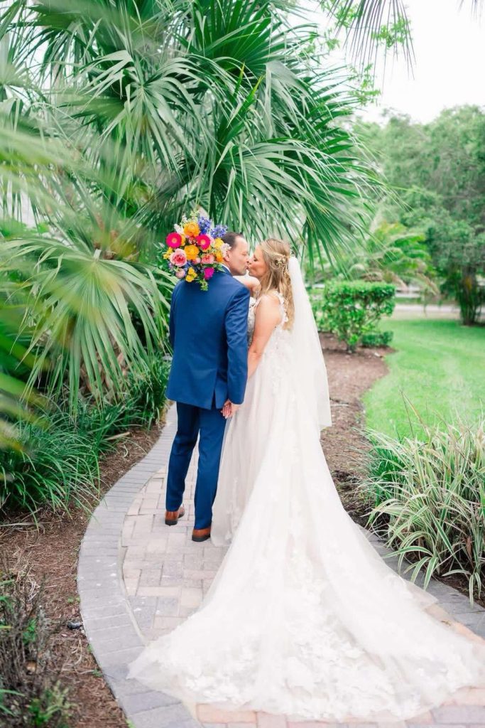 Bride and groom sharing a kiss on a garden pathway surrounded by lush palm trees, with the bride wearing a flowing lace wedding gown and long veil.