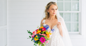 Bride holding a vibrant bouquet of colorful flowers while standing on a white porch in a lace wedding dress and veil.