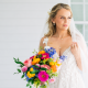 Bride holding a vibrant bouquet of colorful flowers while standing on a white porch in a lace wedding dress and veil.