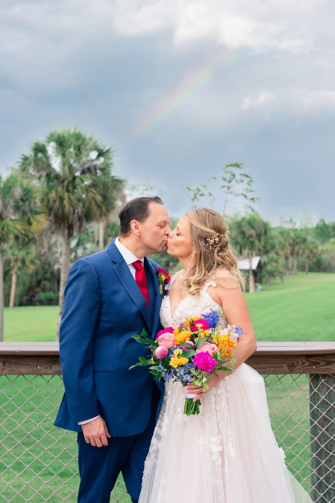 Bride and groom kissing outdoors beneath a rainbow while the bride holds a colorful wildflower-inspired bouquet.