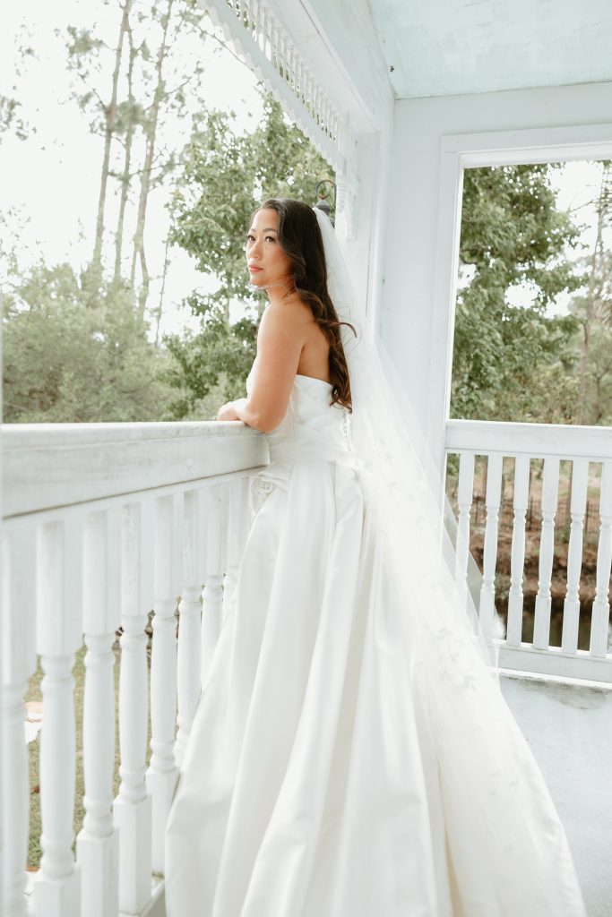 A bride in a strapless white wedding gown and long veil stands on a white porch overlooking a wooded landscape.
