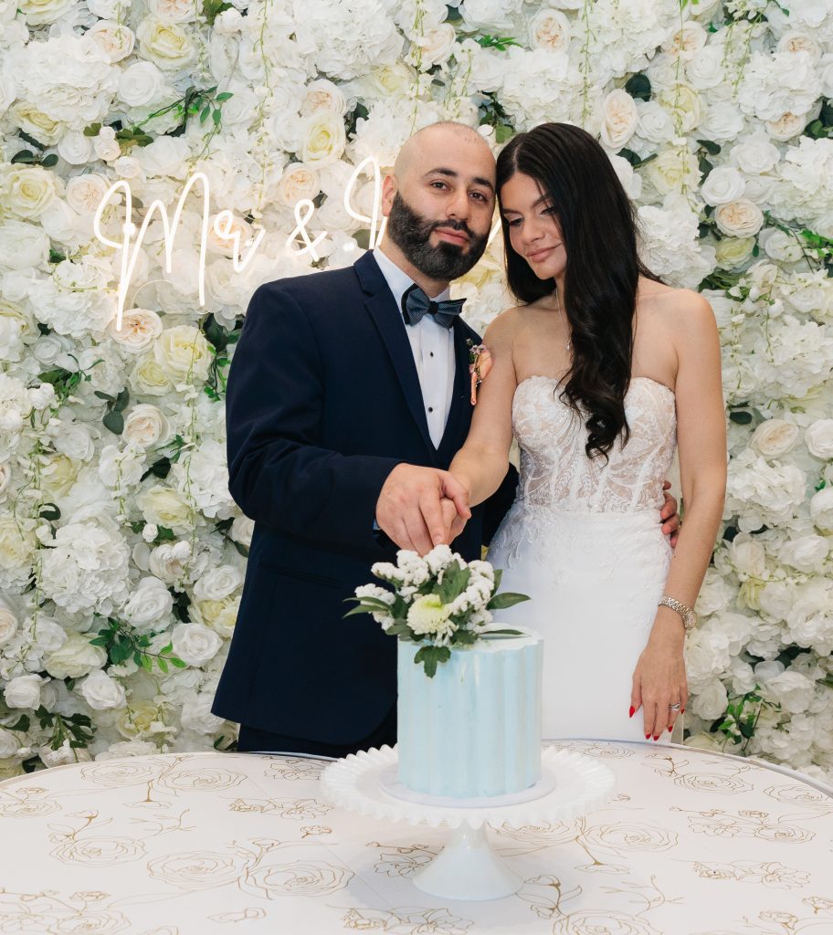 A couple cuts a pale blue wedding cake together in front of a large wall covered with white flowers.